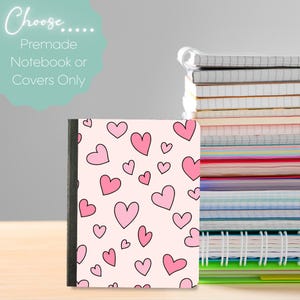 May include: A notebook with a light pink cover featuring a repeating pattern of various sized pink hearts with black outlines. The notebook has a black spine and is positioned next to a stack of spiral-bound notebooks. Text reads: Choose..... Premade Notebook or Covers Only.