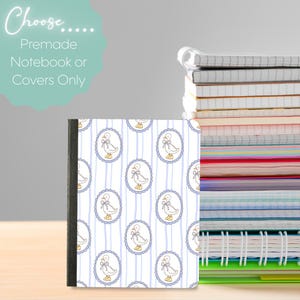 Puede incluir: Un cuaderno con cubierta blanca que presenta un patrón repetitivo de rayas verticales azul claro y marcos ovalados que contienen un pato de dibujos animados con un lazo y botas. El texto Choose Premade Notebook or Covers Only es visible.