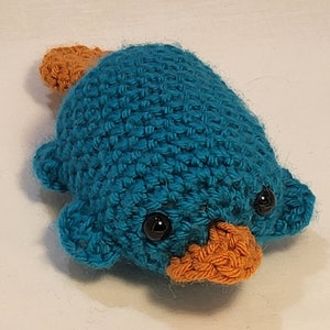 May include: A crocheted plush toy shaped like a platypus. The platypus is blue with orange accents and black eyes.