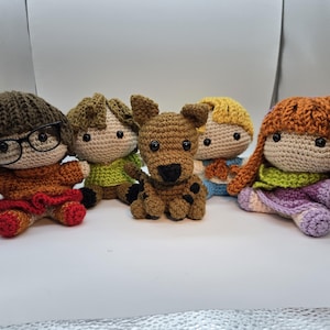 May include: A collection of five handmade crochet dolls, including characters with brown, blonde, and red hair. One doll wears glasses. The dolls are in various outfits, including a brown dog. The dolls are sitting on a white surface.