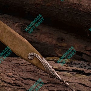 May include: A brown leather knife sheath with silver metal accents and a decorative swirl design. The sheath is resting on a piece of wood.