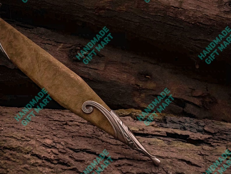 May include: A brown leather knife sheath with silver metal accents and a decorative swirl design. The sheath is resting on a piece of wood.
