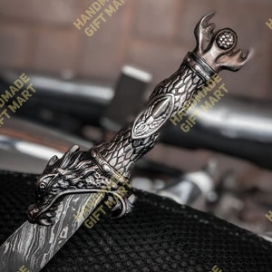 May include: A silver dragon sword with a detailed handle and a textured blade. The sword is resting on a black textured surface. The text "HANDMADE GIFT MART" is visible in the image.