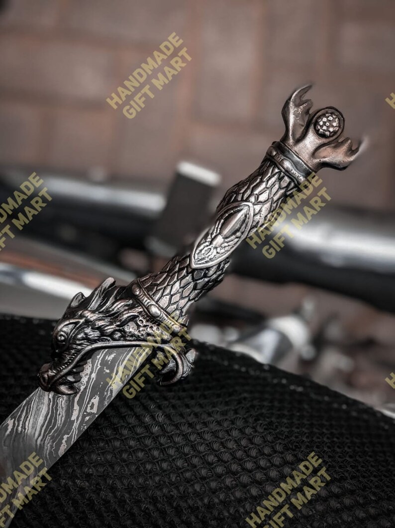 May include: A silver dragon sword with a detailed handle and a textured blade. The sword is resting on a black textured surface. The text "HANDMADE GIFT MART" is visible in the image.