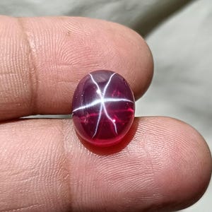 Puede incluir: Una gema pulida, de forma ovalada y color rubí, con un efecto de estrella de seis rayos. La estrella se crea mediante líneas blancas que se cruzan en la superficie de la gema. La gema se sostiene entre los dedos, mostrando su color y claridad.