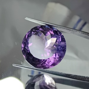 Puede incluir: Una piedra preciosa de amatista redonda y facetada con un tono púrpura. La piedra preciosa está sujeta por unas pinzas.