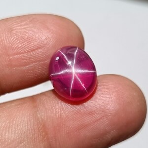 Puede incluir: Una gema de rubí pulida, de forma ovalada, con un efecto de estrella de seis rayos. La gema es de un color rojo rosado intenso y vibrante, con un patrón de estrella blanco brillante que irradia desde el centro. La gema se sostiene entre los dedos.
