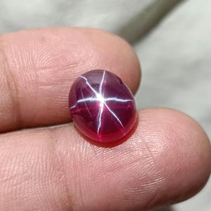 Peut inclure: Une pierre précieuse polie, de forme ovale, rouge foncé, avec un motif en étoile à six rayons. L'étoile est créée par des lignes blanches qui se croisent au centre de la pierre. La gemme est lisse et très brillante.