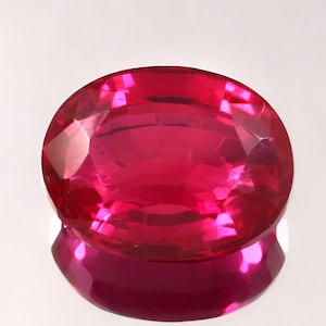 Puede incluir: Una gema ovalada de color rojo intenso y vibrante, con un corte facetado. La gema refleja la luz, mostrando su brillo.