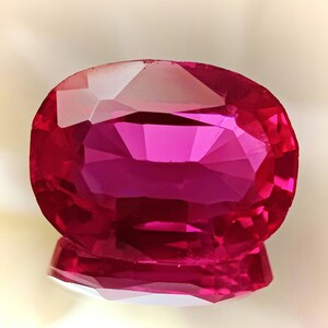 Puede incluir: Una gema ovalada de color rosa vibrante con un corte facetado. La gema refleja la luz, creando un efecto brillante.