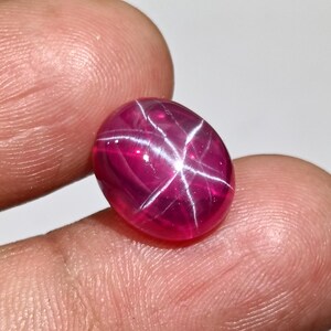 May include: A polished, oval-shaped ruby gemstone with a star effect. The gem is a deep, vibrant red with a six-rayed star pattern across its surface. The stone is held between fingers, showcasing its color and clarity.