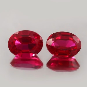 Könnte beinhalten: Zwei oval geschliffene, facettierte Rubin-Edelsteine. Die Rubine sind leuchtend rot und reflektieren das Licht, wodurch ein funkelnder Effekt entsteht. Die Edelsteine werden auf einer reflektierenden Oberfläche präsentiert.
