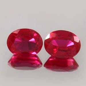 Puede incluir: Dos rubíes facetados de corte ovalado. Las piedras preciosas son de un rojo intenso y vibrante con reflejos internos. Las gemas se exhiben sobre una superficie reflectante, creando una imagen especular de cada piedra.