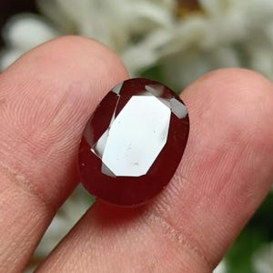 May include: A deep red oval gemstone with a faceted cut. The gemstone is held in a hand.