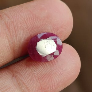 May include: A deep red oval-shaped gemstone with a faceted cut. The gemstone is held in a hand.