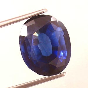 Puede incluir: Una piedra preciosa de zafiro ovalada azul oscuro con un corte facetado. El zafiro está sujeto por unas pinzas.