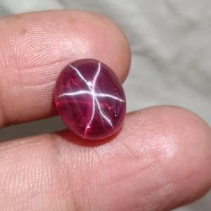Puede incluir: Gema ovalada de color rojo intenso con un patrón de estrella de seis rayos. La superficie pulida de la gema refleja la luz, creando un efecto de estrella. Esta gema es un rubí.
