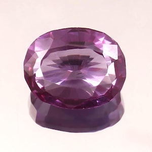May include: A faceted, oval-shaped gemstone in shades of purple. The gem has a polished surface with multiple facets that reflect light, creating a sparkling effect. The gem is set against a reflective surface, enhancing its brilliance.