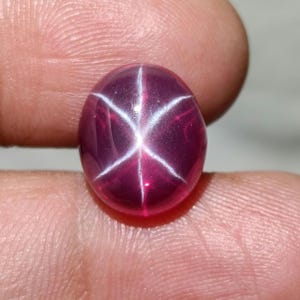 May include: A polished, oval-shaped ruby gemstone with a six-rayed star effect. The gem is a deep, rich red color with a bright white star pattern across its surface. The stone is held between fingers, showcasing its size and luster.