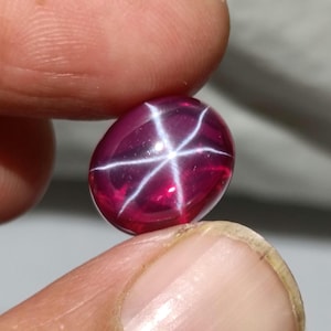 May include: A polished, oval-shaped ruby gemstone. The deep red gem features a six-rayed star pattern, with white lines radiating from the center. The gem is held between two fingers, showcasing its size and shape.