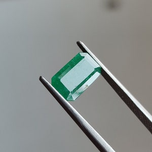 May include: A green emerald gemstone held by tweezers. The emerald is cut into a rectangular shape with facets.