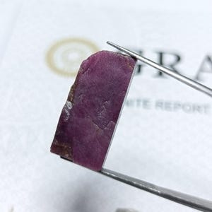 Könnte beinhalten: Ein roher, ungeschliffener Rubin-Edelstein mit tiefmagentafarbener Tönung. Der Rubin wird von einer Pinzette gehalten, die seine raue, natürliche Textur und Form zeigt. Der Hintergrund enthält einen weißen Bericht.
