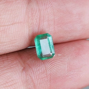 May include: A vibrant green emerald gemstone with a rectangular cut. The emerald is set in a clear, faceted setting.