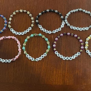 May include: A collection of ten beaded bracelets with various colors and designs. Each bracelet features white letter beads spelling out words like "Karma", "Evermore", "Ready For It", and "August". The bracelets are arranged on a brown surface.