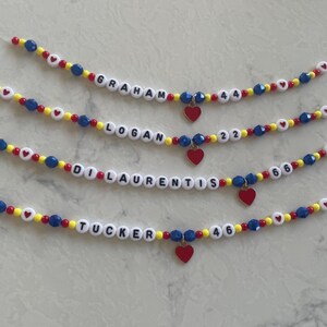 May include: Four beaded bracelets featuring white letter beads spelling names like Graham, Logan, Di Laurentis, and Tucker, alongside number beads 44, 22, 66, and 46. The strands include red, yellow, and blue beads, small white heart beads, and a red heart charm.