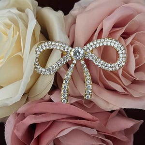 May include: A gold-toned bow brooch encrusted with clear rhinestones, centered with a larger stone. The brooch is set against a backdrop of cream and pink roses, creating a romantic and elegant aesthetic.