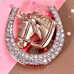 May include: A rose gold-colored horse head brooch set within a horseshoe shape. The horseshoe and horse head are embellished with clear, round rhinestones. The brooch is a decorative accessory.