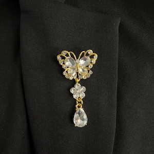 May include: A gold-toned butterfly brooch with clear crystal accents. The butterfly design is embellished with small crystals and features a dangling flower and teardrop-shaped crystal pendant. The brooch is pinned to a black fabric.