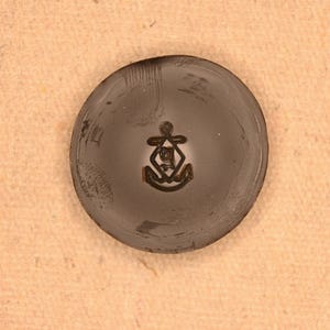 May include: A dark gray, circular object with a raised emblem featuring an anchor and a stylized design. The object has a slightly textured surface and appears to be made of metal. The emblem is centered on the object.