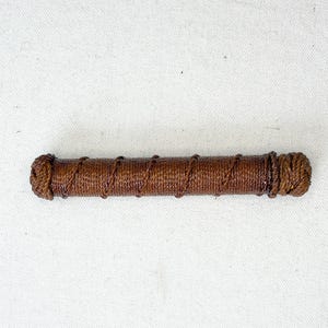 May include: A cylindrical, brown, woven object with tightly wrapped rope detailing. The ends are thicker and more textured, suggesting a handle or decorative element. The item is set against a plain white background.