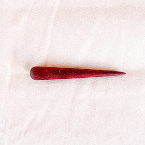 May include: A handcrafted, tapered awl with a rounded handle. The awl is made of dark red wood, with visible grain, and is designed for piercing or marking materials. The tool is set against a white fabric background.