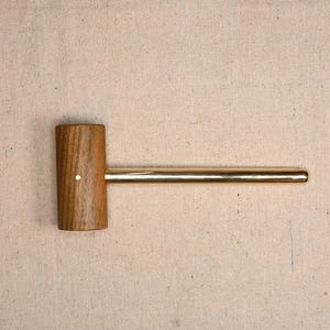 May include: A wooden mallet with a cylindrical head and a long, brass handle. The mallet head is made of a light brown wood and has a small, white dot on its side. The handle is a shiny gold color.