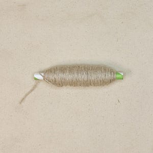 May include: A spool of natural-colored twine, tightly wound around a white cardboard tube with green accents. A loose end of the twine extends from the spool. The background is a light beige fabric.