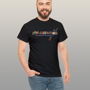 May include: Black t-shirt with the word "IMAGINATION" in colorful letters. The phrase "is creating my reality" is below the main word. The design includes colorful dots and shapes. The shirt is worn by a person.