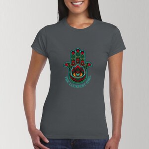 Könnte beinhalten: Ein dunkelgraues T-Shirt mit einem farbenfrohen Hamsa-Hand-Design und dem Text "The Luckiest Girl" in Türkis.