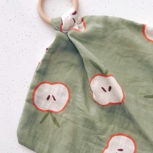 May include: A green baby security blanket with a wooden ring. The blanket features a repeating pattern of white apple halves with red outlines and green stems. The ring is made of light-colored wood.