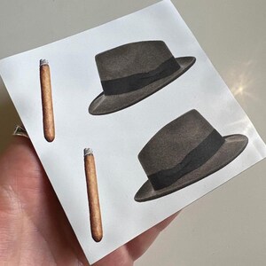 May include: Two black fedora hats with black bands and two brown cigars on a white background.