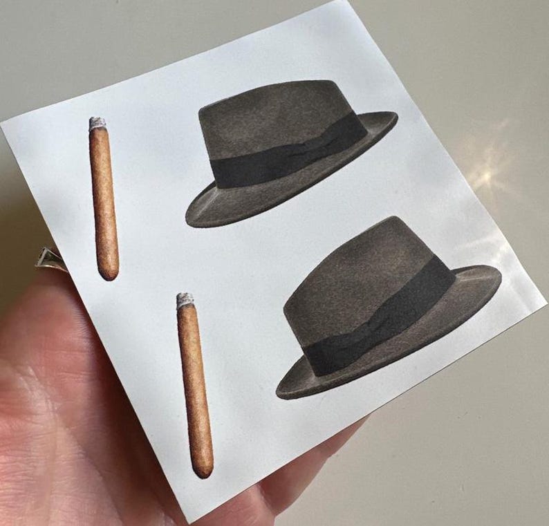 May include: Two black fedora hats with black bands and two brown cigars on a white background.