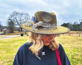 May include: Olive green fedora hat with a wide brim, adorned with a leopard print ribbon and a decorative bow. The hat features subtle floral embellishments and is set against a blurred outdoor background.