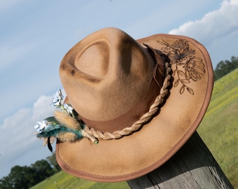 May include: A brown felt fedora hat with a braided rope band and a floral design etched into the brim. The hat is decorated with a small bouquet of white flowers and a blue feather.
