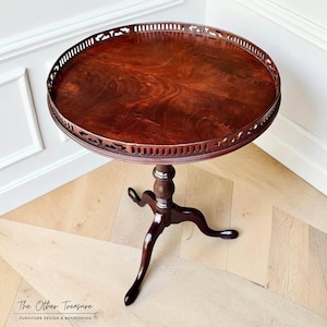 May include: A round, dark brown wooden pedestal table featuring a circular top with a decorative, pierced gallery railing around the edge. The table is supported by a central turned column that branches into three curved legs with feet.