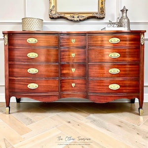 May include: A mahogany wood dresser featuring a serpentine front with four drawers on each side and four central drawers. The piece includes oval brass pulls, small shield-shaped escutcheons, and gold-toned accents on the fluted side columns and feet.