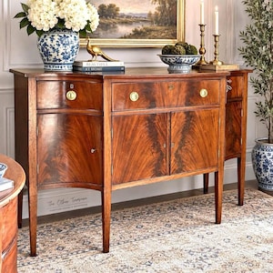 May include: A mahogany sideboard featuring a curved front, two central cabinet doors, and two side drawers with brass hardware. The piece has tapered legs and a polished wood grain finish, displaying a classic furniture design with keyholes.