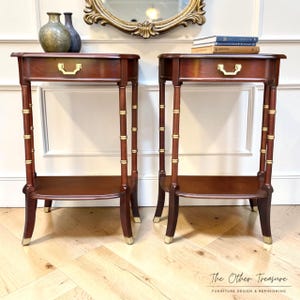 May include: Two matching mahogany-toned side tables with a drawer and lower shelf. The legs have a bamboo-like design with gold accents. Each table has a gold-toned drawer pull and gold feet. The tables are displayed in front of a white wall.
