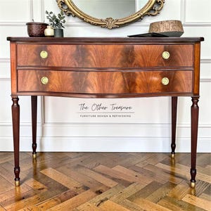 May include: A mahogany-toned wooden console table with two drawers and brass hardware. The table has a curved front and slender, fluted legs. The text "The Other Treasure" is visible on the front of the table.