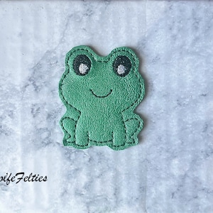May include: Green felt frog applique with black stitching and white eyes. The frog is sitting with its legs tucked under its body.
